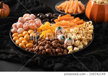 Halloween sweet charcuterie board with pumpkin, popcorn, orange chocolate candy, spider, crackers on black background. Scary decoration. Festive food for kids party. Close up. Halloween sweet charcuterie board with pumpkin, popcorn, orange chocolate candy, spider, crackers on black background. Scary decoration. Festive food for kids party. Close up. 130742149