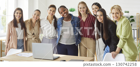 Smiling diverse women standing by desk in office hugging and laughing, showing unity and teamwork. Smiling diverse women standing by desk in office hugging and laughing, showing unity and teamwork. 130742258