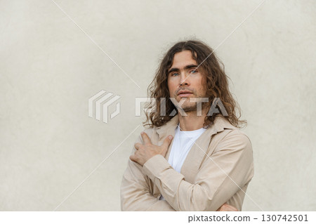 Handsome caucasian man, brunette with curly long hair wearing beige casual attire stands against neutral background looking at camera. Contemplation and introspection. Space for design. 130742501