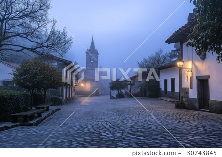 Mystical atmosphere enveloping a quaint European village square at dawn, with fog hovering over the cobblestone streets and a church steeple piercing the sky 130743845