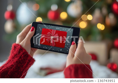Woman holding a tablet displaying a gift card with a red ribbon, in front of a decorated Christmas tree 130743951