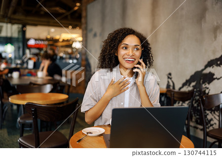 A Joyful Woman Engaging in a Phone Conversation while Sitting in a Cozy Cafe with her Laptop 130744115
