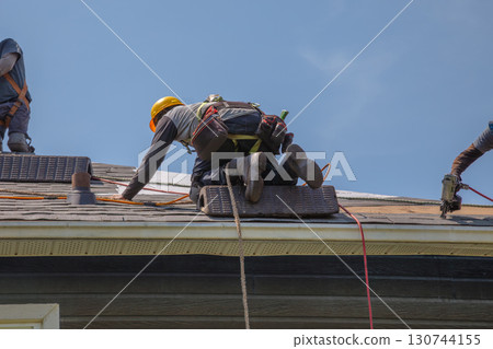 Construction Workers with Safety Equipment Installing Asphalt Shingles on Residential Roof Construction Workers with Safety Equipment Installing Asphalt Shingles on Residential Roof 130744155