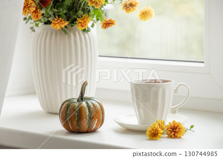 Autumn tea in white cup, decorative pumpkin and season flowers on white window sill. Cozy home interior and calm. Close up. Autumn tea in white cup, decorative pumpkin and season flowers on white window sill. Cozy home interior and calm. Close up. 130744985