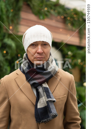 Portrait of serious and lonely man wearing white beanie and cozy scarf, with blurred Christmas festive background. Cold winter weather. Xmas holiday. Portrait of serious and lonely man wearing white beanie and cozy scarf, with blurred Christmas festive background. Cold winter weather. Xmas holiday. 130744989