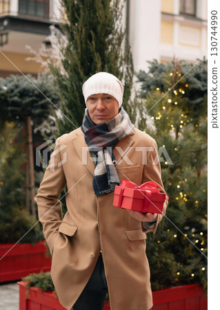Man wearing beige coat giving Christmas red gift box near Xmas tree at city. Xmas shopping and happy holiday. Outdoors. Close up. Man wearing beige coat giving Christmas red gift box near Xmas tree at city. Xmas shopping and happy holiday. Outdoors. Close up. 130744990