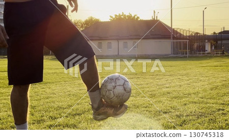 Male feet of professional footballer kicking ball at green field. Legs of young man juggling soccer ball on stadium at sunset. Sportsman practicing tricks outdoor. Freestyle football concept 130745318