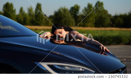 Beautiful smiling girl bought new electrical vehicle and hugging hood auto. Happy young woman enjoying modern electric automobile. Concept of buy or rent car. Slow motion 130745332