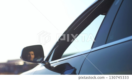 Young smiling businessman in shirt leaning out of driver window at modern electric car and looking into camera. Portrait of handsome successful entrepreneur in innovative electrical vehicle. Slow mo 130745333