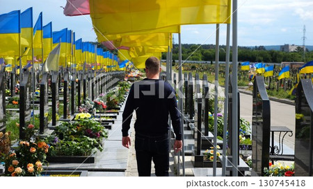 Mourning man go among countless graves of ukrainian soldiers with national flags at military cemetery in Kharkiv. Scene reflects personal sorrow and nation collective loss caused by russian aggression 130745418
