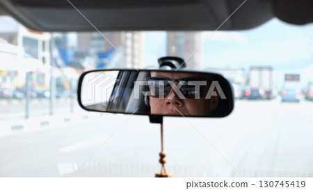 Eyes in sunglasses of male driver reflecting in rearview mirror while riding motor auto. Young guy looking concentrated to urban road during operating vehicle. Confident man driving modern car at city Eyes in sunglasses of male driver reflecting in rearview mirror while riding motor auto. Young guy looking concentrated to urban road during operating vehicle. Confident man driving modern car at city 130745419