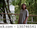 A thoughtful woman in a grey oversized T-shirt stands on a rainy balcony surrounded by green foliage and white architecture 130746541
