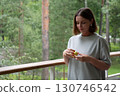 Young woman with short hair and red nails gently holds a green apple while standing on a porch during a summer rain in the forest 130746542
