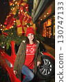 a pretty young woman in a red hat with a bubo smiles against the backdrop of a red retro car with Christmas gifts on the roof, Christmas lights and snow. 130747133