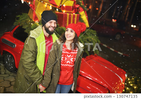 a couple of lovers - a man and a woman hold hands and smile against the background of a red retro car with Christmas gifts on the roof 130747135