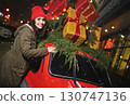 a pretty young woman in a red hat with a bubo smiles against the backdrop of a red retro car with Christmas gifts on the roof, Christmas lights and snow. 130747136