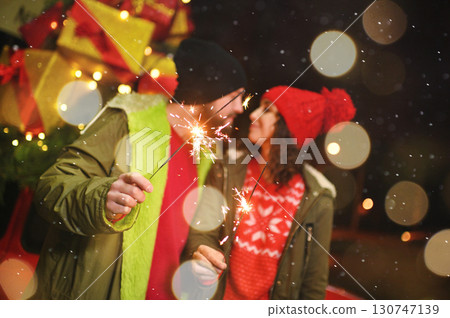 a couple of lovers - a man and a woman kiss against the backdrop of Christmas gifts and snowfall, holding sparklers in their hands 130747139