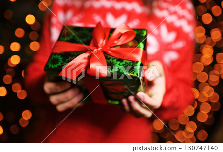 close-up - a girl in a red sweater with a Scandinavian pattern holds a Christmas present in her hands against the background of Christmas lights. 130747140
