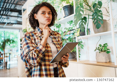 A contemplative woman in a modern workspace is thoughtfully engaging with her tablet device 130747162