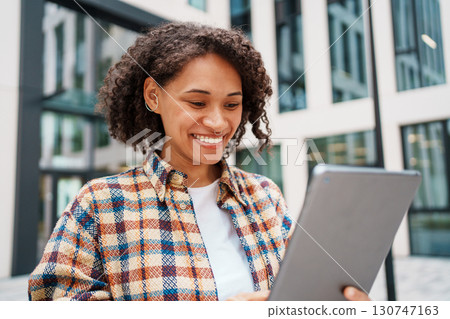 A Smiling Woman Happily Using a Tablet Outdoors in a Modern Urban Setting Surrounded by Nature 130747163