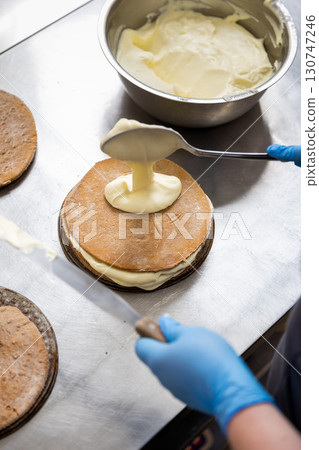 Pastry chef spreading cream on cake layer in professional bakery kitchen, close-up Pastry chef spreading cream on cake layer in professional bakery kitchen, close-up 130747246