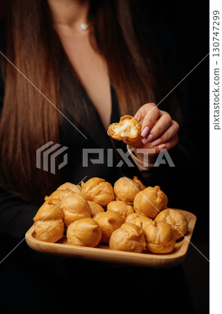 Woman holding a bitten cream puff over a tray of profiteroles in elegant setting 130747299