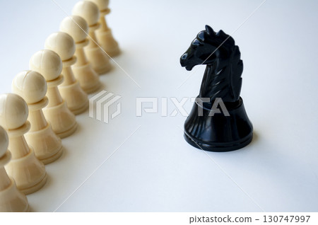 A single black knight chess piece placed prominently in front of several beige pawns aligned in formation symbolizing strategy and direction. 130747997