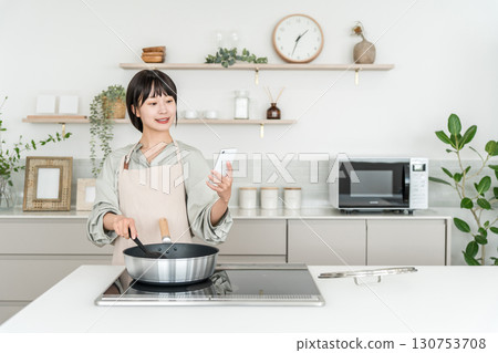 A young Asian woman wearing an apron cooking in the kitchen while looking at her smartphone 130753708