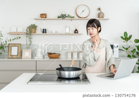 A young Asian woman wearing an apron cooking while looking at a laptop in the kitchen 130754176