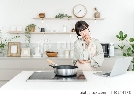 A young Asian woman in an apron cooking while looking at a laptop in the kitchen (thinking, worrying, dislikes, hates) 130754178
