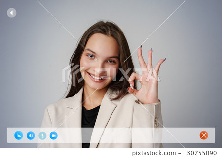 Young woman in a light suit making an okay gesture in a professional setting against a neutral background Young woman in a light suit making an okay gesture in a professional setting against a neutral background 130755090