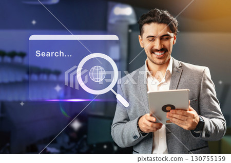 Man using tablet for online research in a modern workspace with digital interface displayed in the background during daylight Man using tablet for online research in a modern workspace with digital interface displayed in the background during daylight 130755159