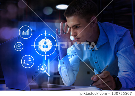 Young man focused on digital interface while working on laptop in modern office during evening hours 130755507