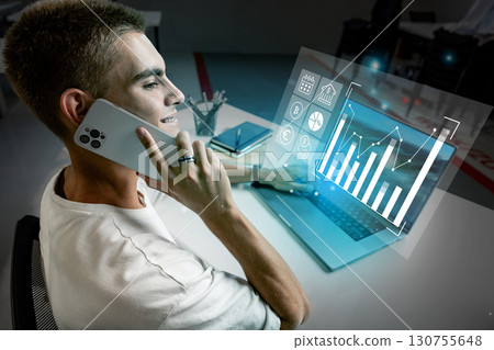 Young man talking on smartphone while analyzing financial data on laptop in modern workspace 130755648