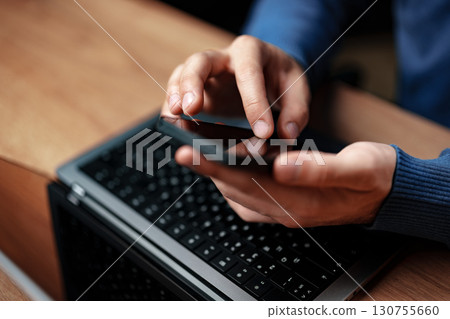 Man using smartphone while working on laptop at a wooden desk in a cozy indoor setting during daytime Man using smartphone while working on laptop at a wooden desk in a cozy indoor setting during daytime 130755660