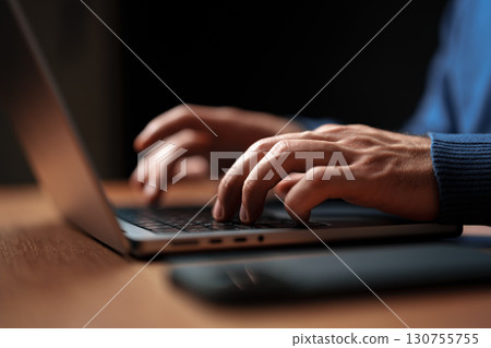 Hands typing on a laptop in a dimly lit workspace during evening hours 130755755