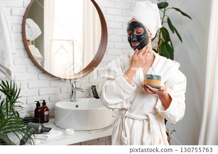 Woman applying face mask while looking in mirror in a stylish bathroom setting during morning routine Woman applying face mask while looking in mirror in a stylish bathroom setting during morning routine 130755763