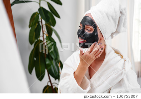 Woman applying charcoal face mask while enjoying self-care routine at home in bright living room Woman applying charcoal face mask while enjoying self-care routine at home in bright living room 130755807