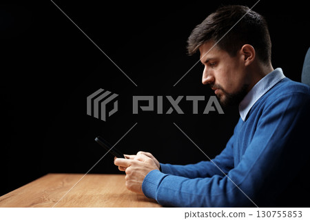 Man using smartphone while sitting at desk in a dimly lit room during evening hours 130755853