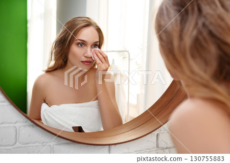 Woman applying skincare in front of round mirror in a bright bathroom after showering and wrapping her hair in a towel 130755883