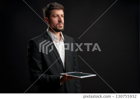 Professional man in formal attire holds tablet while looking thoughtfully against dark background Professional man in formal attire holds tablet while looking thoughtfully against dark background 130755932
