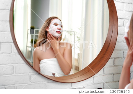 Enjoying self-care with a charcoal face mask in a bright, modern bathroom while admiring reflections in the mirror 130756145