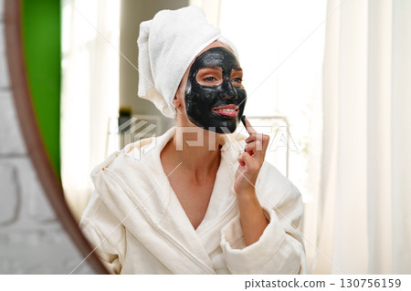 Woman applying black facial mask while looking in mirror at home on a relaxing afternoon Woman applying black facial mask while looking in mirror at home on a relaxing afternoon 130756159