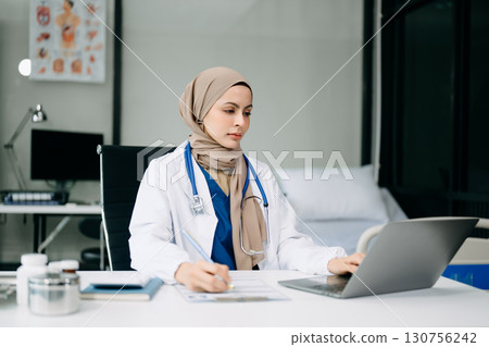 Beautiful physician doctor sit on table in office hospital. Attractive therapist female practitioner pharmacist smile and use tablet and laptop. 130756242