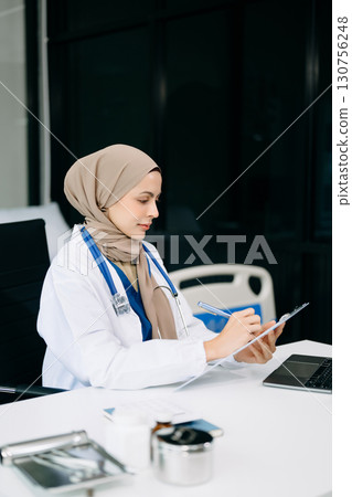 Beautiful physician doctor sit on table in office hospital. Attractive therapist female practitioner pharmacist smile and use tablet and laptop. 130756248