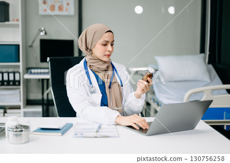 Beautiful physician doctor sit on table in office hospital. Attractive therapist female practitioner pharmacist smile and use tablet and laptop. 130756258