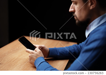 Man focused on smartphone at wooden table in dimly lit indoor space during evening hours Man focused on smartphone at wooden table in dimly lit indoor space during evening hours 130756263