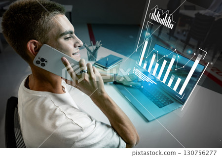 Young professional working on a laptop and talking on the phone in a modern office setting with data visualization 130756277