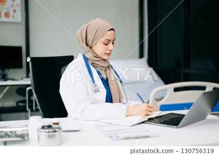 Beautiful physician doctor sit on table in office hospital. Attractive therapist female practitioner pharmacist smile and use tablet and laptop. 130756319