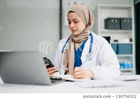 Beautiful physician doctor sit on table in office hospital. Attractive therapist female practitioner pharmacist smile and use tablet and laptop. 130756322